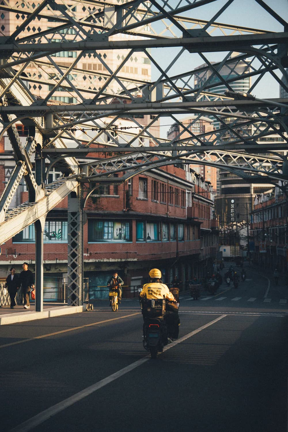 Residents and delivery riders moving beneath a historic steel bridge in Shanghai