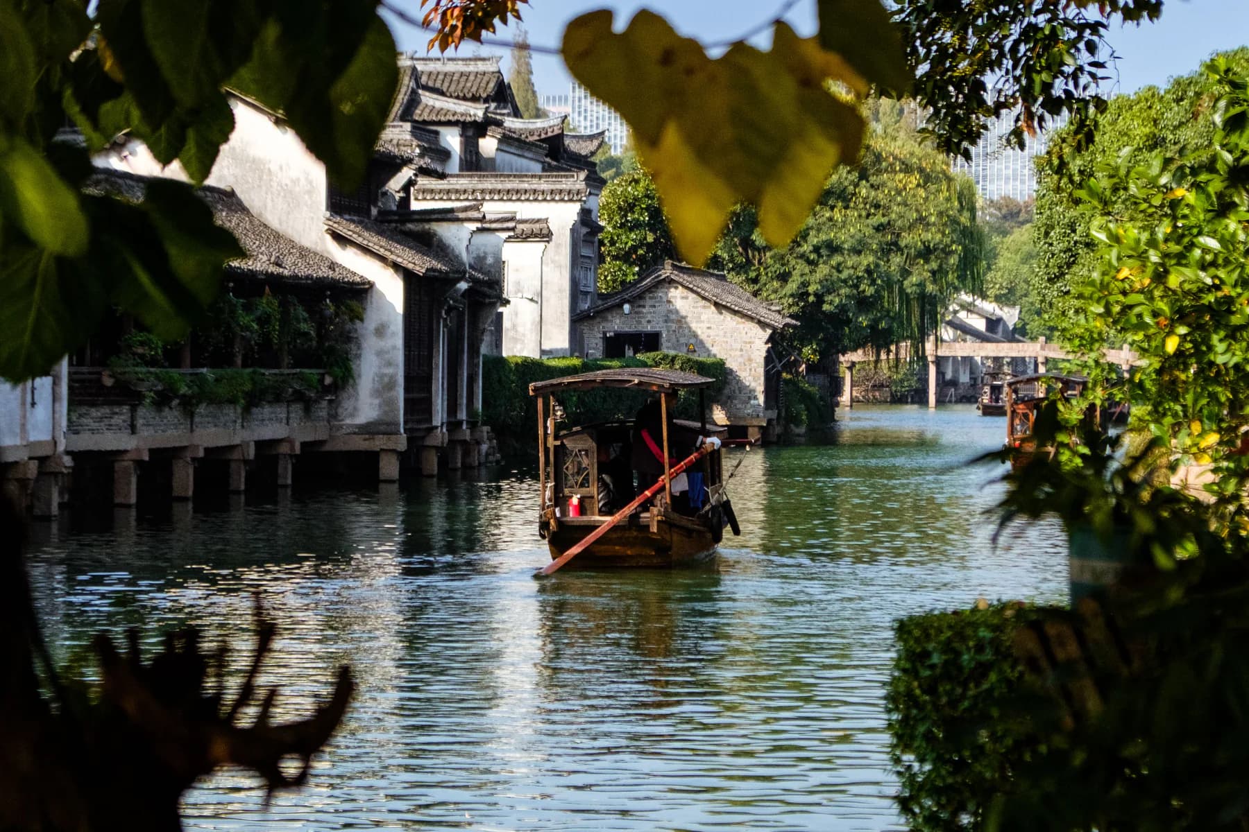 A small boat moving along a green Suzhou canal between whitewashed houses and leafy banks